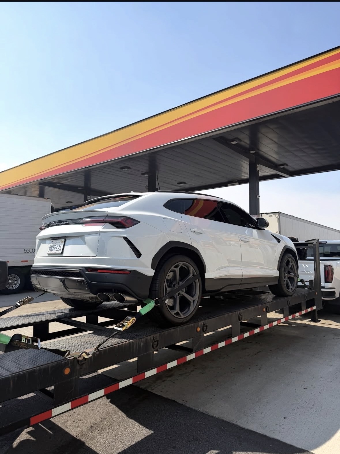 White Lamborghini Urus secured on a Royalty Auto Transport flatbed at a gas station